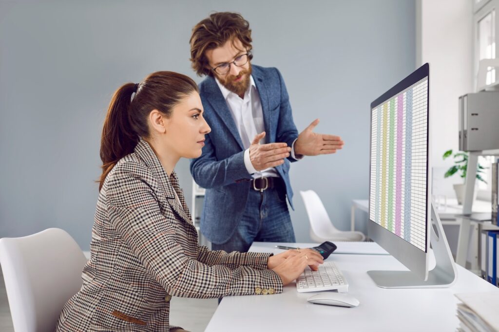 People working in office. Male manager talking to female trainee, giving advice and explaining something. Woman sitting at desk, looking at spreadsheets on computer screen and listening to colleague